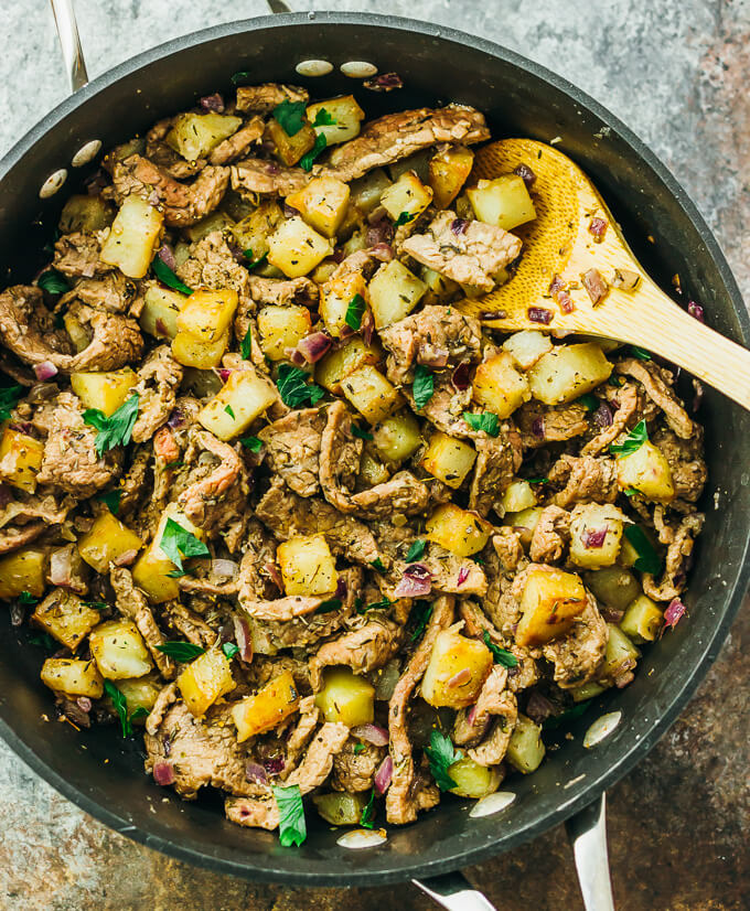A large skillet with thinly sliced steak, cubed potatoes, and onions