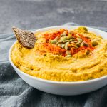 Pumpkin hummus plated in a serving bowl topped with pumpkin seeds, paprika, and flax seed cracker