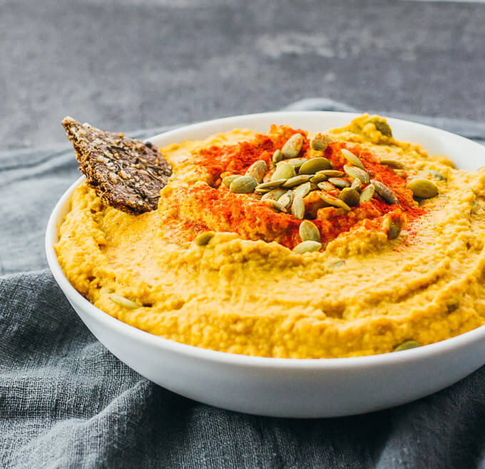 Pumpkin hummus plated in a serving bowl topped with pumpkin seeds, paprika, and flax seed cracker