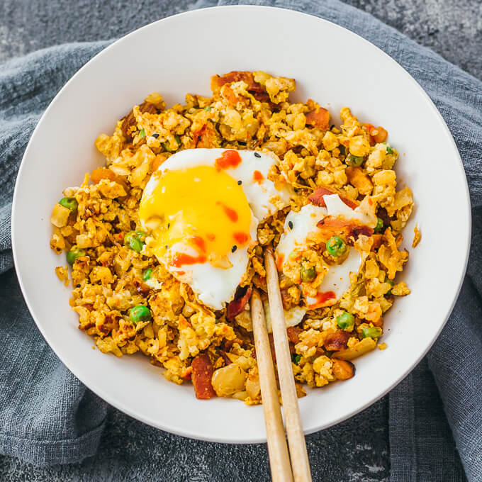 Bacon kimchi cauliflower fried rice served in a white bowl and topped with fried egg and sriracha